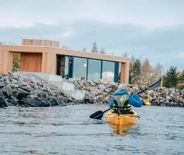 En person i gul kajakk padler mot et moderne bygg i tre ved vannkanten omgitt av steiner.