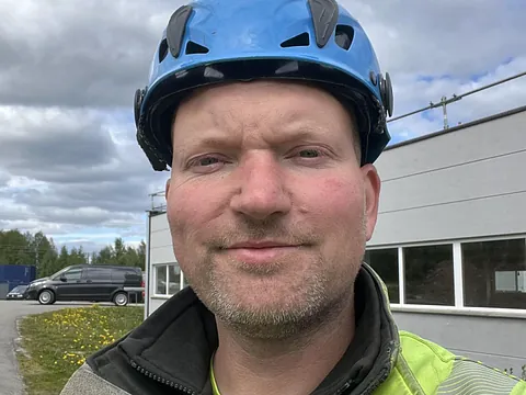 Mann i gul arbeidsjakke og blå hjelm tar selfie ute foran grå bygning og skyet himmel.