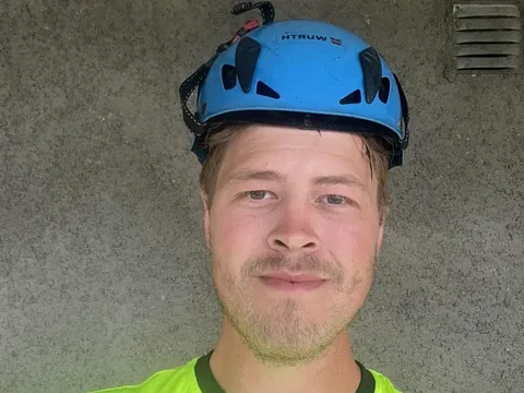 Ung mann med blå hjelm og neongul trøye tar en selfie foran en betongvegg og bølgeblikk-tak.
