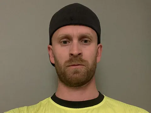 Mann i gul t-skjorte og bakvendt caps står innendørs foran en grå vegg og ser rett i kamera.